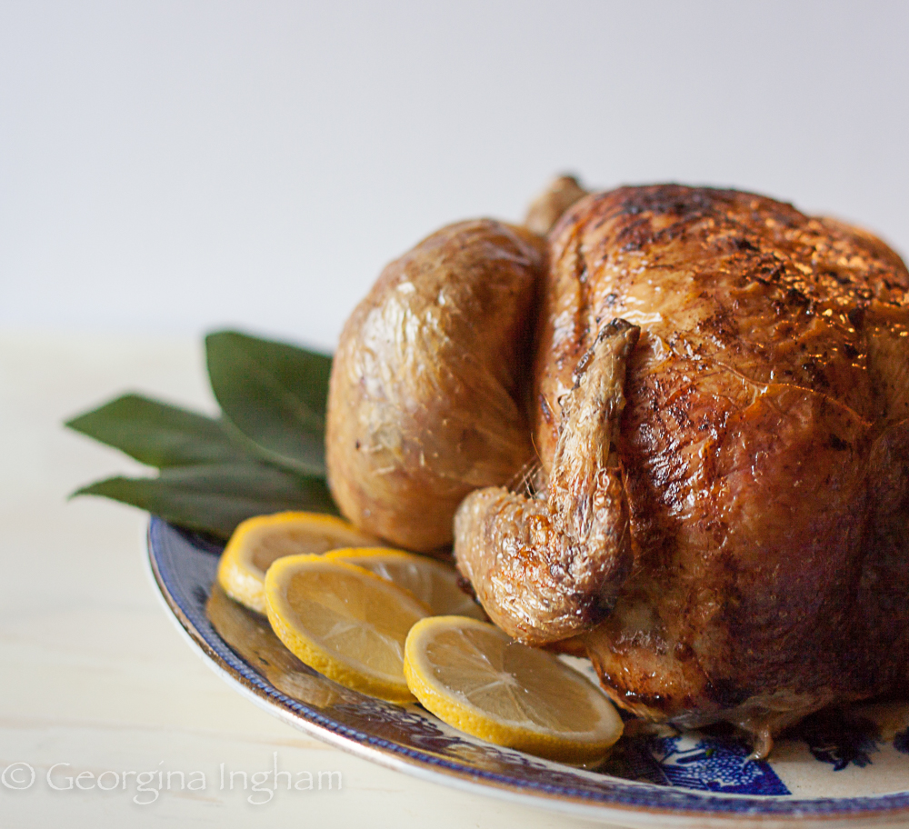 Golden lemon and herb roast chicken with bay leaves and sliced lemon, ready to serve on a blue patterned platter.